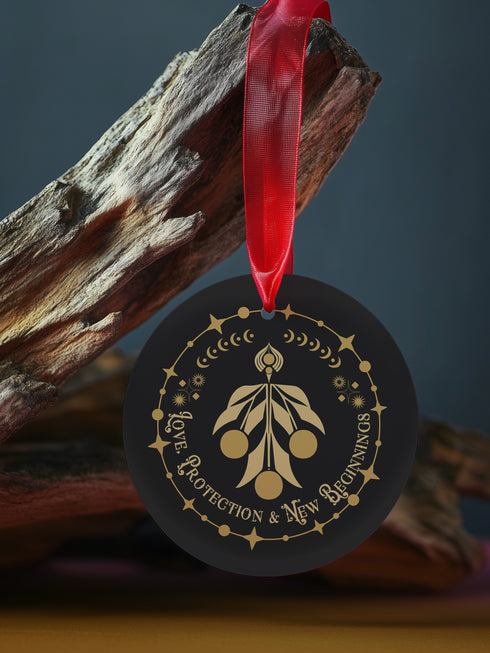 Round metal ornament featuring golden mistletoe design with Art Deco border incorporating stars and moon phases. 3.5-inch diameter piece with "Love, Protection & New Beginnings" text and crimson ribbon. Double-sided design on black background.